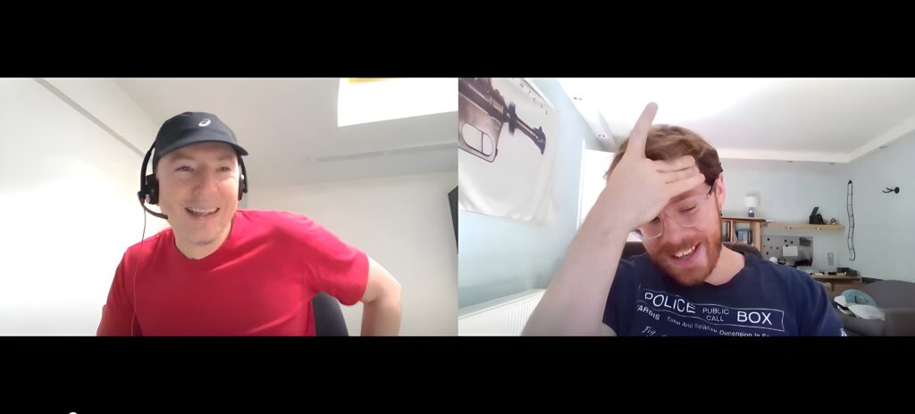 Two men have a video call; the one on the left wears a headset and smiles, while the one on the right holds his forehead and looks amused or exasperated.