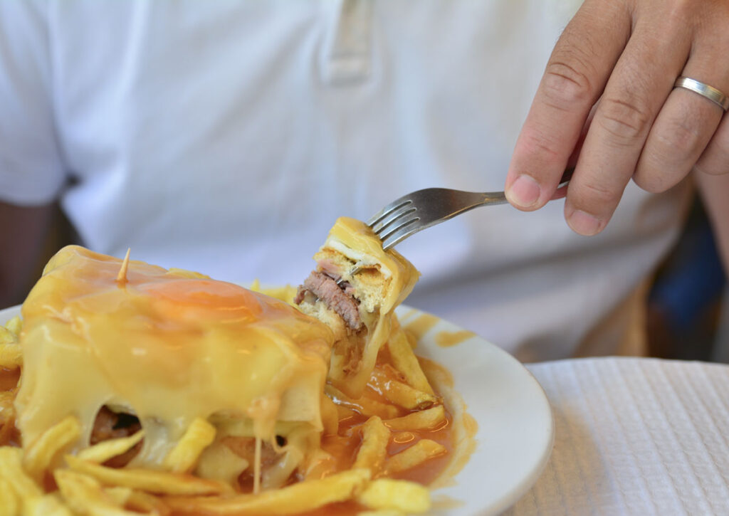 A person wearing a white shirt uses a fork to pick up a bite of a sandwich topped with melted cheese and sauce, served with fries on a white plate.